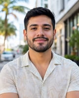 Man with dark hair and beard smiling at camera wearing light beige short-sleeve shirt on urban street with palm trees