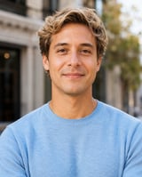 Portrait of a man with wavy blonde hair wearing a light blue sweater against an urban street background